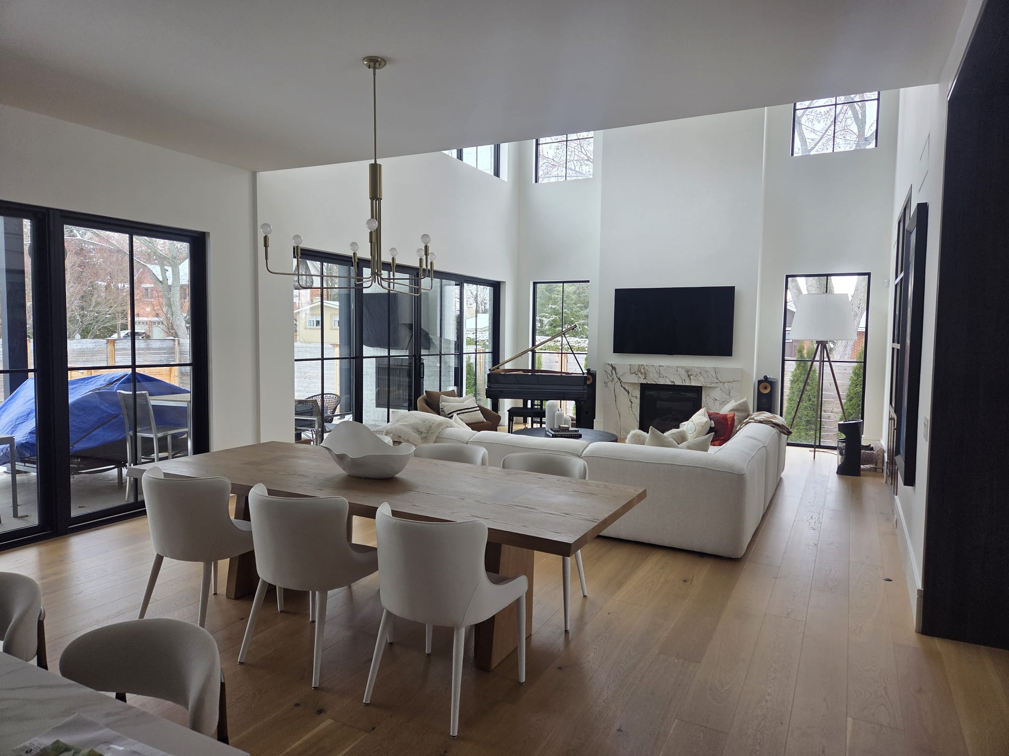 Luxury open-concept living and dining room renovation in Oakville featuring large black-framed windows, light wood floors, a modern chandelier, and a grand piano.